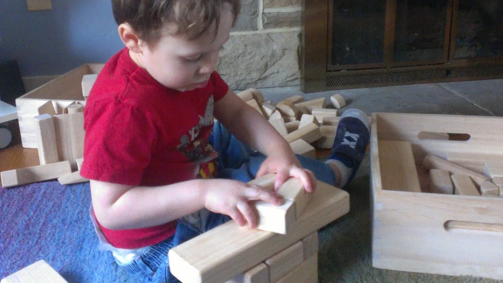 Young child deeply absorbed in screen-free play, building with wooden unit blocks on a living room rug